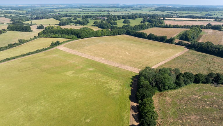 A high-angle drone photograph showing a patchwork of agricultural fields and open countryside. Large rectangular fields in shades of green and golden brown are bordered by hedgerows and clusters of mature trees. A dirt track curves around the edge of a harvested field in the foreground, while rolling farmland stretches to the horizon beneath a clear blue sky.